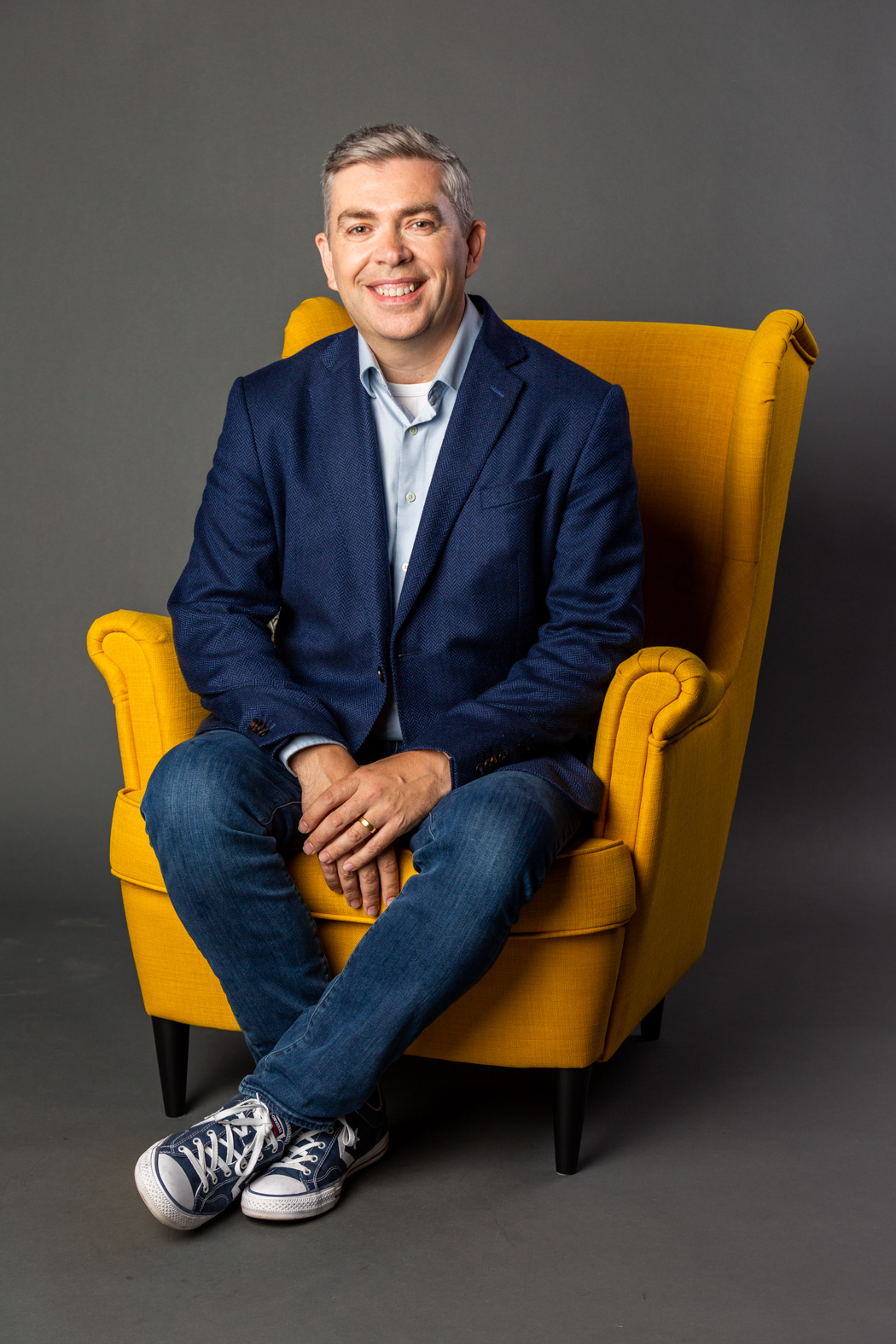 Andrew Finlayson seated on a yellow chair, in a blue shirt and jacket.